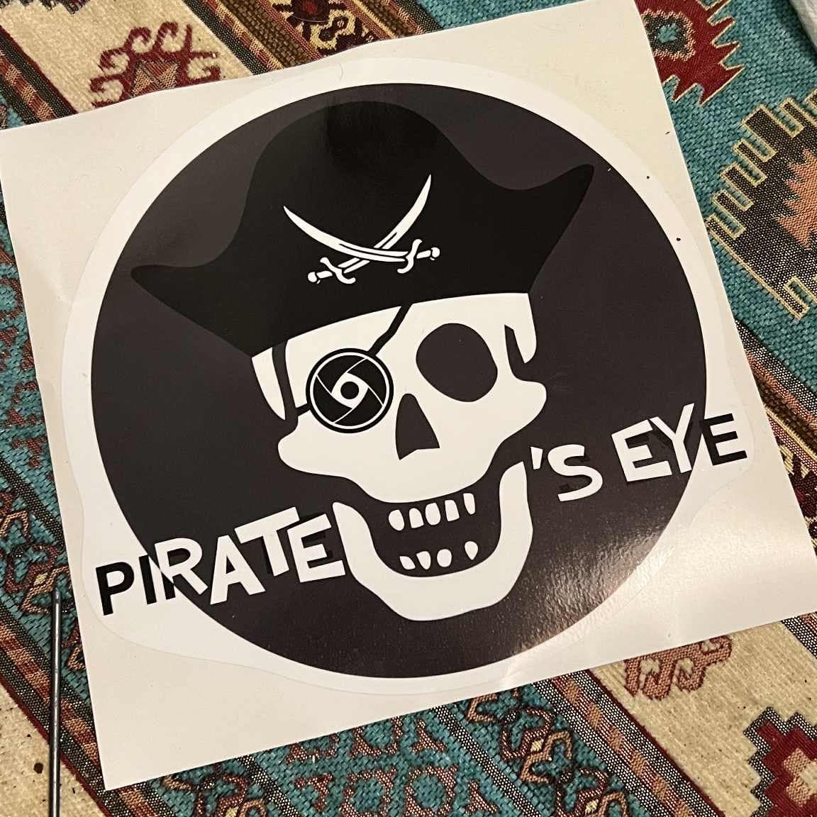 A large square Pirate's Eye sticker lying on a patterned rug: a white skull in a black tricorn hat, one eye replaced by a camera shutter, on a black disc.